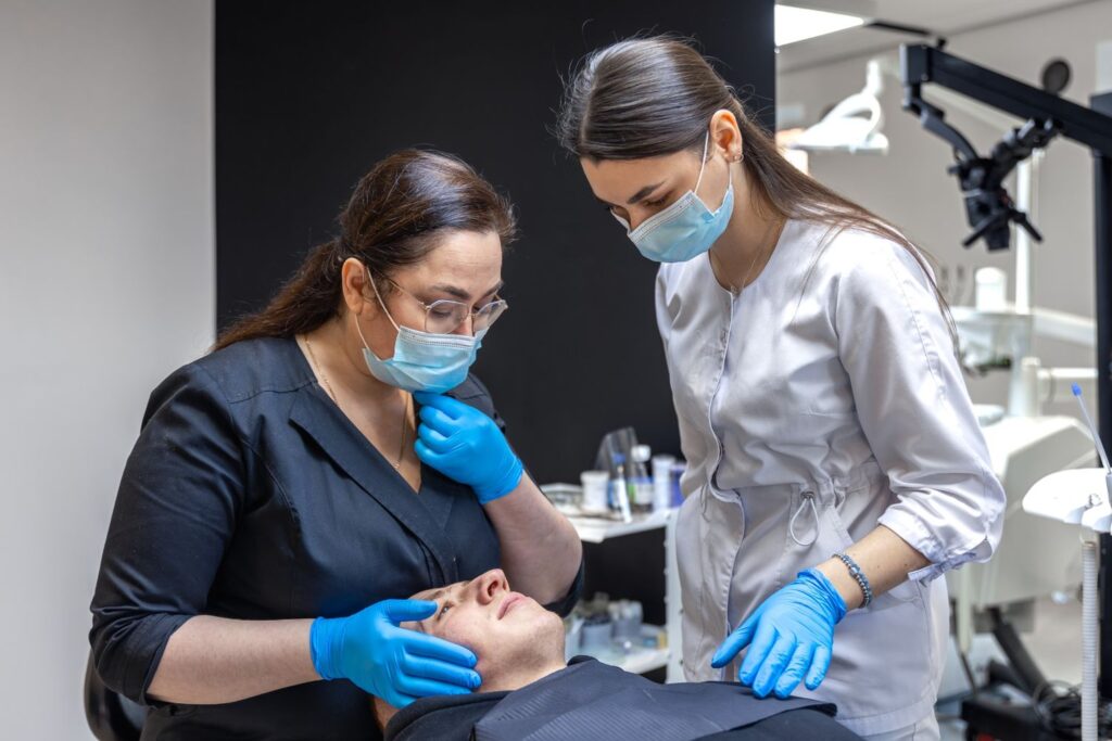 Two female dermatology professionals examining a patient’s facial skin during a clinical consultation, wearing masks and gloves in a medical setting.
